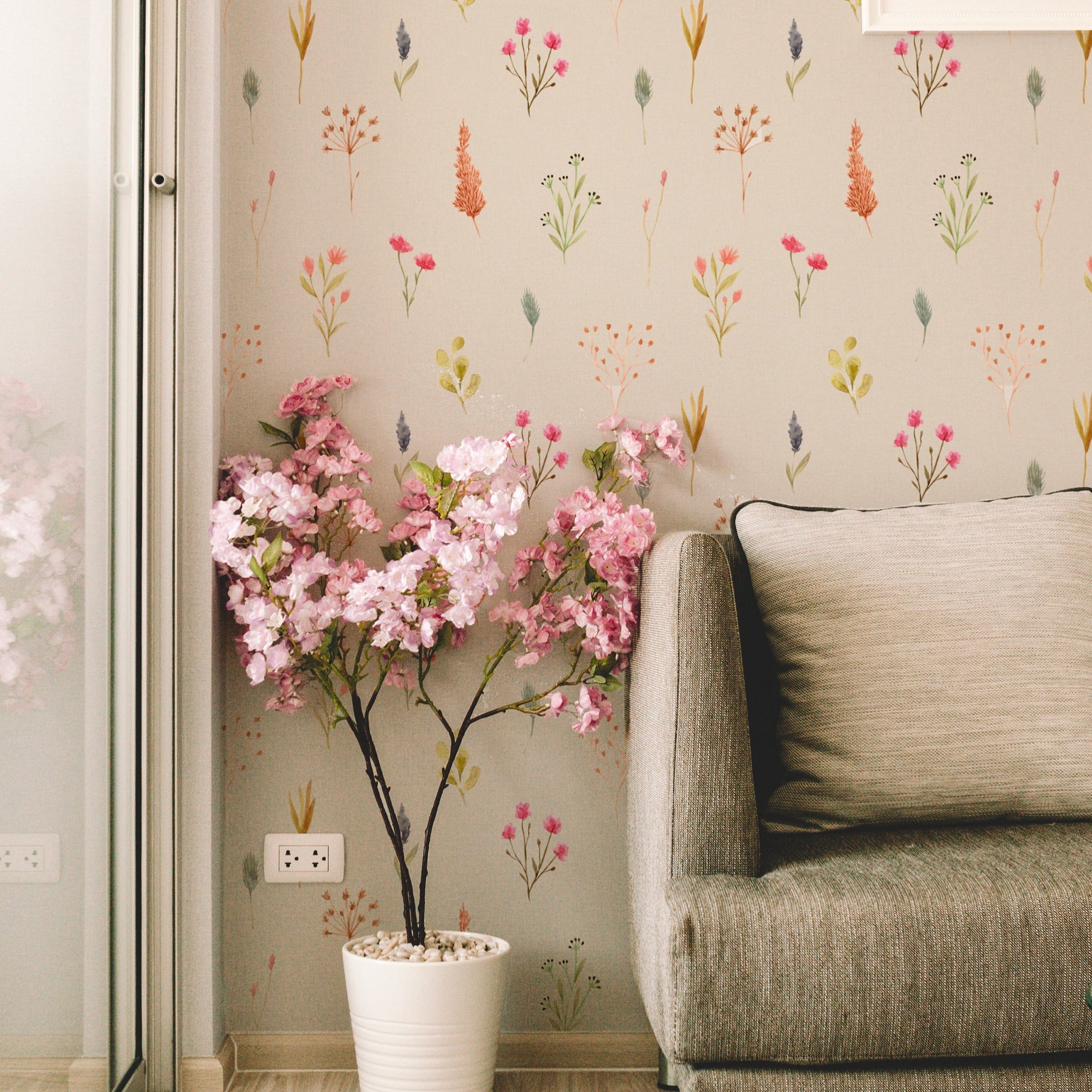 A stylish living area where a plush grey sofa is set against the Watercolour Floral Wallpaper II, with vibrant pink blossoms in a white pot complementing the wallpaper's floral motif, enhancing the room's elegance and botanical charm.