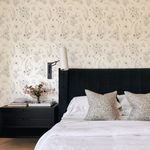A chic bedroom design where the 'Wildflower Sketch Wallpaper' provides a soft and artistic backdrop to a modern black nightstand, an upholstered black headboard, and decorative elements including a white cylindrical lamp and a vase with pink flowers.