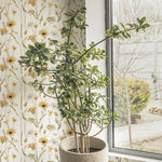A potted green plant sits beside a window, with the Watercolour Sunflower Wallpaper in the background. The sunlight illuminates the wallpaper’s soft watercolor sunflowers and wildflowers, creating a serene indoor garden atmosphere.