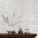 An interior setup showcasing the Painted Abstract Wallpaper applied to a wall. The abstract, free-form hazelnut lines weave across a white backdrop, complementing the rustic wooden console table in the foreground. On the table, a white ribbed vase with dried lavender, a vintage brass mister, and a sculptural white candle holder add to the organic aesthetic of the scene.