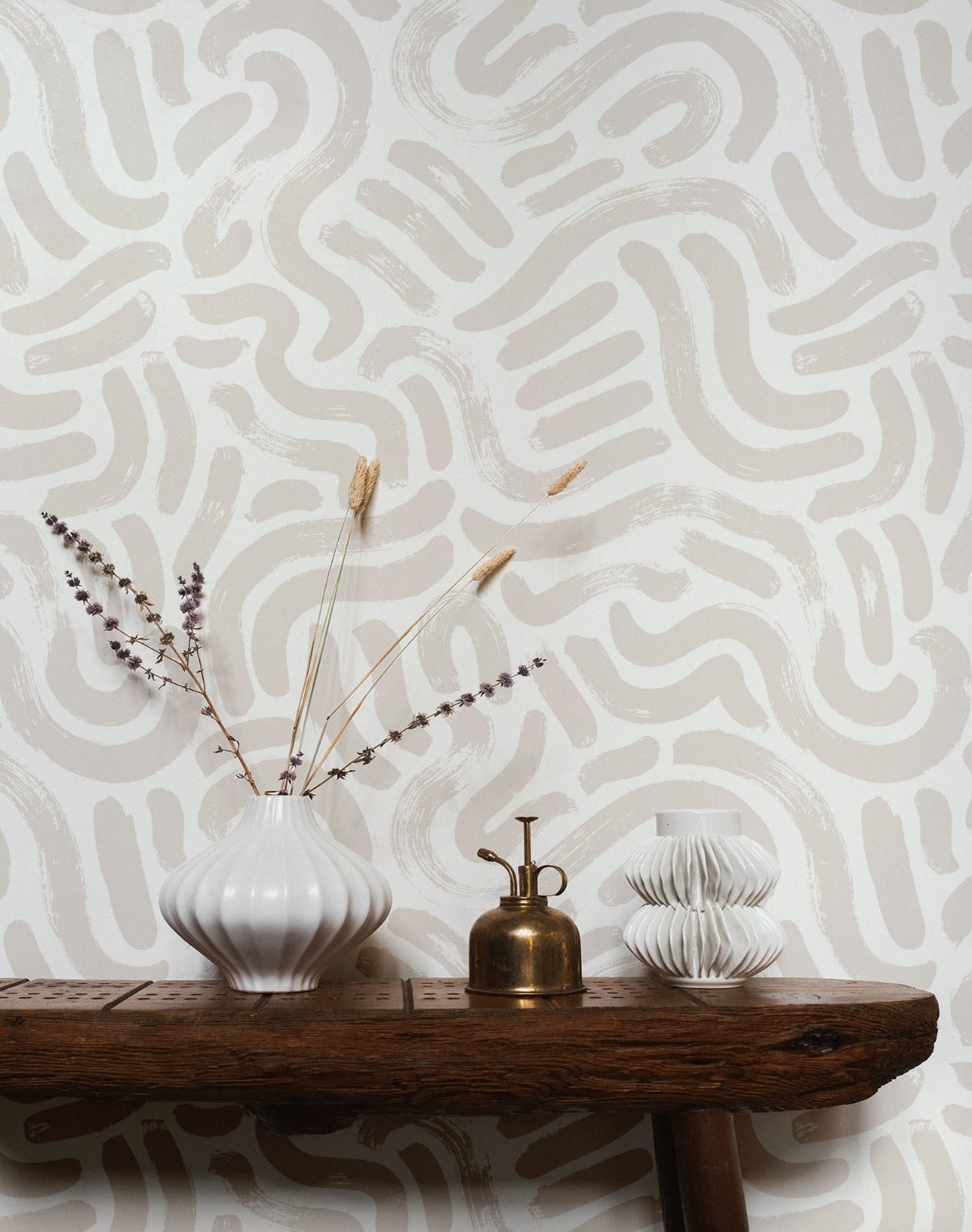 An interior setup showcasing the Painted Abstract Wallpaper applied to a wall. The abstract, free-form hazelnut lines weave across a white backdrop, complementing the rustic wooden console table in the foreground. On the table, a white ribbed vase with dried lavender, a vintage brass mister, and a sculptural white candle holder add to the organic aesthetic of the scene.