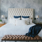 An elegant bedroom featuring a tufted cream headboard against the Pale Blue Brush Stroke Wallpaper, which adds depth and a tranquil feel to the room. The bedding is adorned with white and navy blue pillows, complemented by a bench at the foot of the bed, enhancing the room's sophisticated and restful atmosphere.