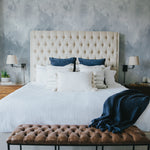 An elegant bedroom featuring a tufted cream headboard against the Pale Blue Brush Stroke Wallpaper, which adds depth and a tranquil feel to the room. The bedding is adorned with white and navy blue pillows, complemented by a bench at the foot of the bed, enhancing the room's sophisticated and restful atmosphere.