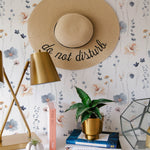 A chic workspace with a straw hat hanging on the wall covered in Watercolour Floral - Summer wallpaper, complemented by a gold desk lamp and a potted plant, creating a tranquil home office.