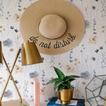 A chic workspace with a straw hat hanging on the wall covered in Watercolour Floral - Summer wallpaper, complemented by a gold desk lamp and a potted plant, creating a tranquil home office.