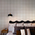 An elegant bedroom interior where the Traditional Tartan Plaid Wallpaper adds a subtle yet impactful backdrop to the stylish monochrome bedding and dark, curvaceous headboard, harmoniously paired with a modern black pendant light and wooden nightstand.
