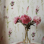A corner of a room showing a portion of the 'Blooming Spring Wallpaper' with a small round table holding a clear glass vase of vibrant pink peonies. The floral arrangement complements the pastel pink flowers and green leaves on the wallpaper.