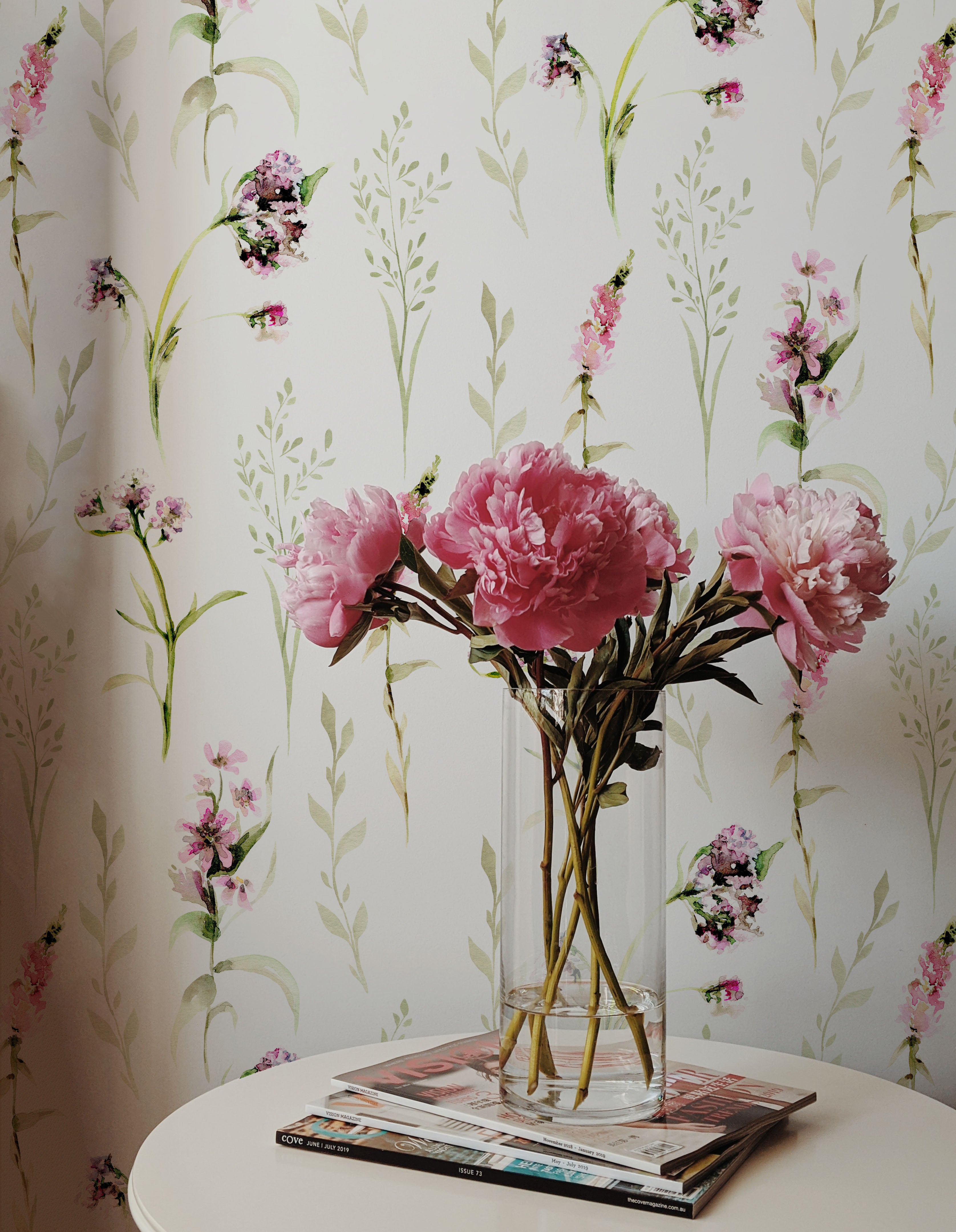 A corner of a room showing a portion of the 'Blooming Spring Wallpaper' with a small round table holding a clear glass vase of vibrant pink peonies. The floral arrangement complements the pastel pink flowers and green leaves on the wallpaper.