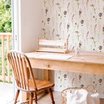 Interior view of a cozy workspace with a wooden desk and chair set against a wall covered in 'Blooming Spring Wallpaper.' The wallpaper features an elegant pattern of pink flowers and green leaves. Natural light streams in from a window to the left, overlooking a green outdoor area.