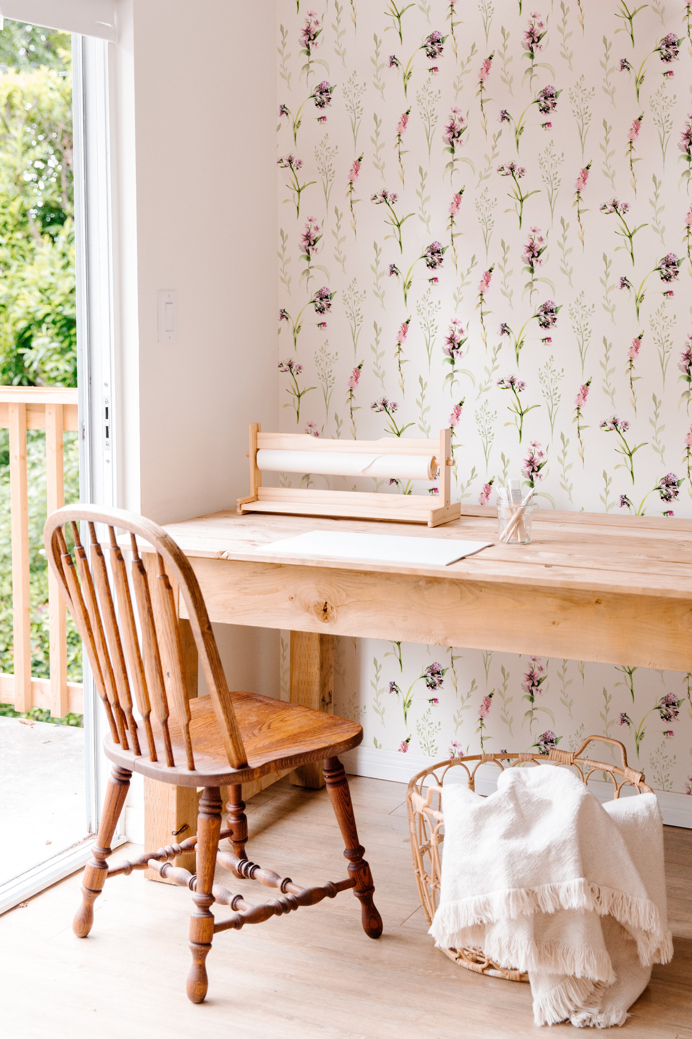 Interior view of a cozy workspace with a wooden desk and chair set against a wall covered in 'Blooming Spring Wallpaper.' The wallpaper features an elegant pattern of pink flowers and green leaves. Natural light streams in from a window to the left, overlooking a green outdoor area.