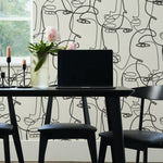 A modern dining area featuring Minimalist Faces Wallpaper with a black line art pattern of abstract faces on a white background. The space includes a black table, black chairs, a vase with flowers, and black candleholders.