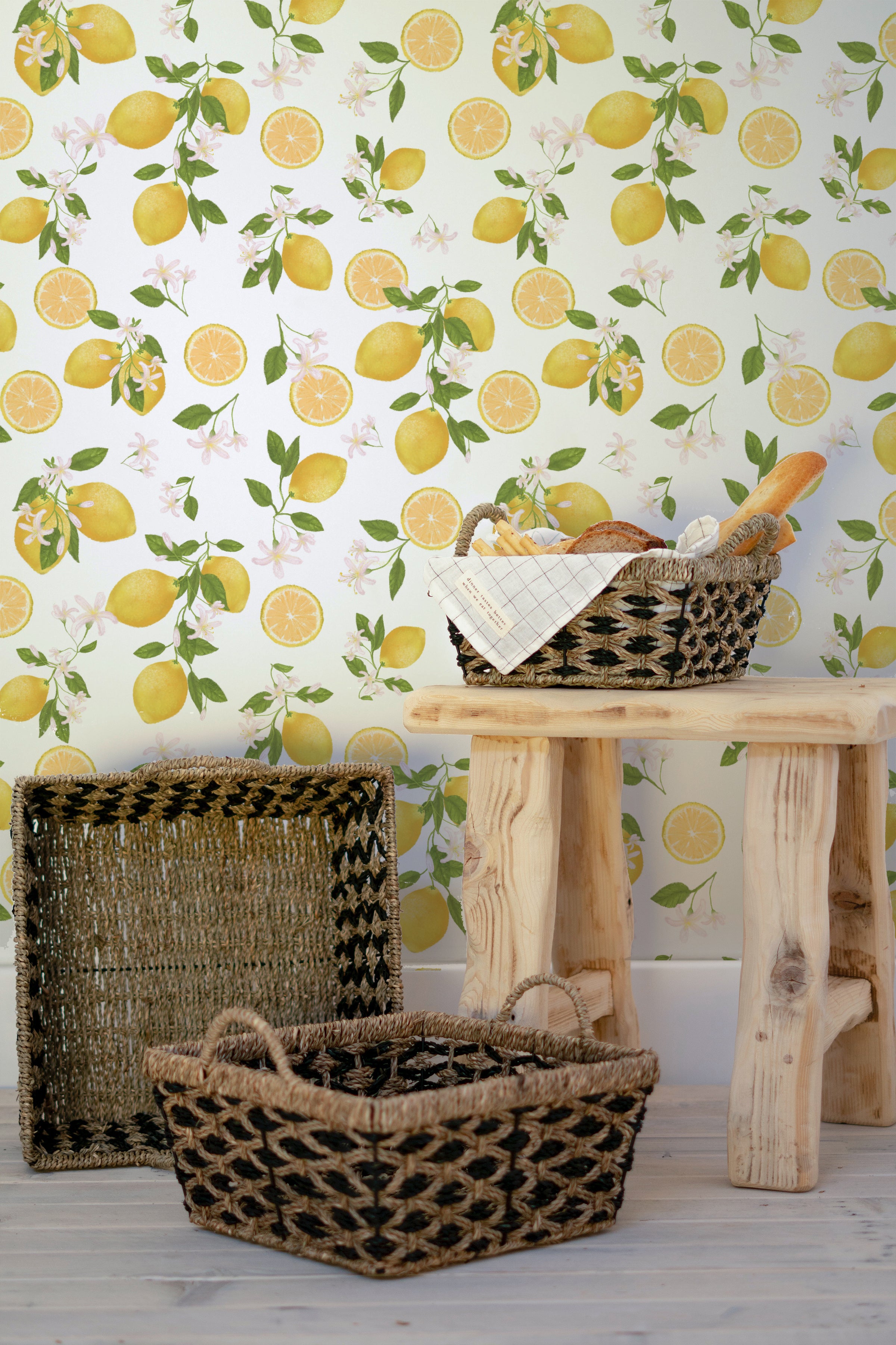 Lemon Floral Wallpaper with a bright and cheerful pattern of yellow lemons, green leaves, and white flowers, enhancing a wall behind a rustic wooden table and woven baskets
