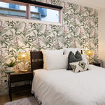 A cozy bedroom with a bed and nightstands against a wall covered in tropical branches wallpaper. The wallpaper features an intricate design of green leaves and pinkish-red flowers on a light background, adding a vibrant and refreshing touch to the room. The nightstands are adorned with lamps and potted plants, enhancing the serene ambiance.