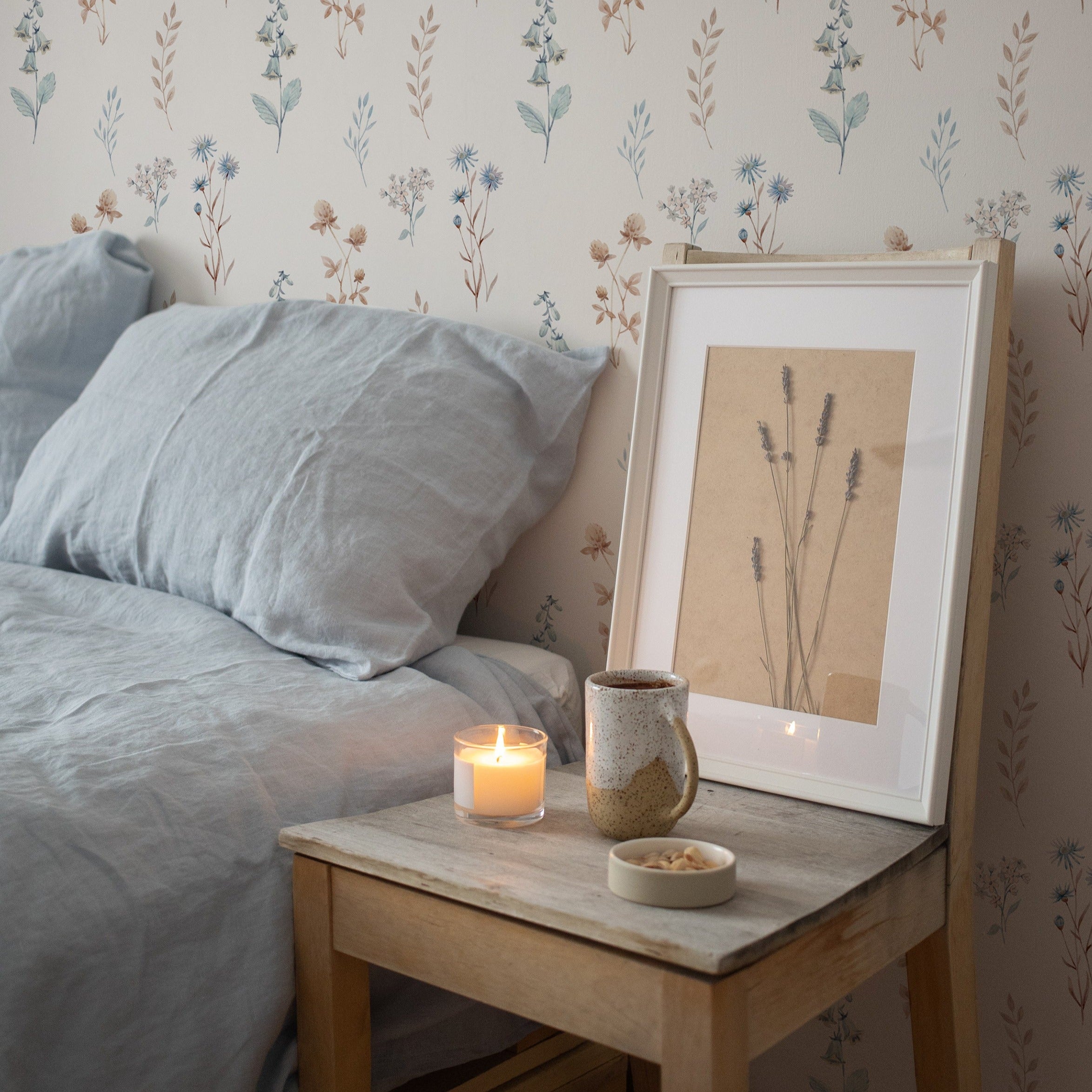 A bedroom setting showcasing the Bluebell Floral Wallpaper as a backdrop. The décor includes a comfy bed with grey linens, a wooden bedside table with a framed picture, a ceramic mug, and a lit candle, creating a relaxing ambiance.