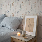 A bedroom setting showcasing the Bluebell Floral Wallpaper as a backdrop. The décor includes a comfy bed with grey linens, a wooden bedside table with a framed picture, a ceramic mug, and a lit candle, creating a relaxing ambiance.
