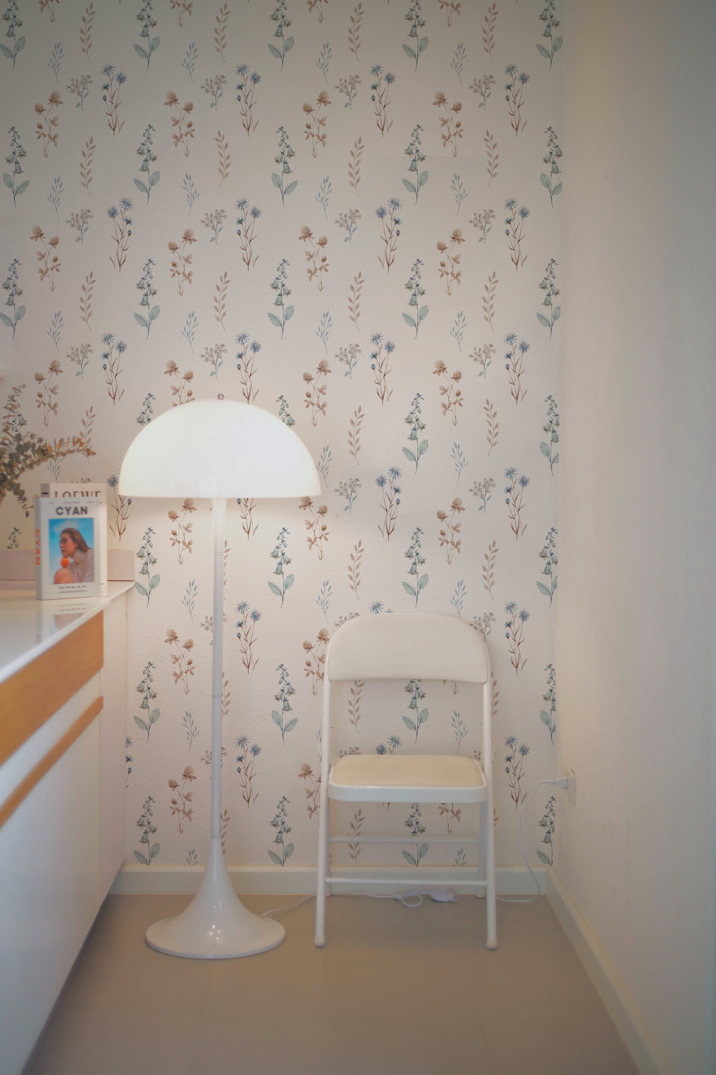 A cozy corner featuring Bluebell Floral Wallpaper with delicate botanical patterns in shades of blue, beige, and green. The scene includes a stylish white floor lamp and a chair, enhancing a serene, light-filled room.
