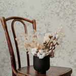 An aged wooden chair set against a wall covered in 'Rose Bouquet Wallpaper,' which showcases a detailed pattern of roses and foliage in muted grey on a light background. Atop the chair is a black vase filled with a mix of dried flowers and cotton branches, complementing the vintage aesthetic of the setting.