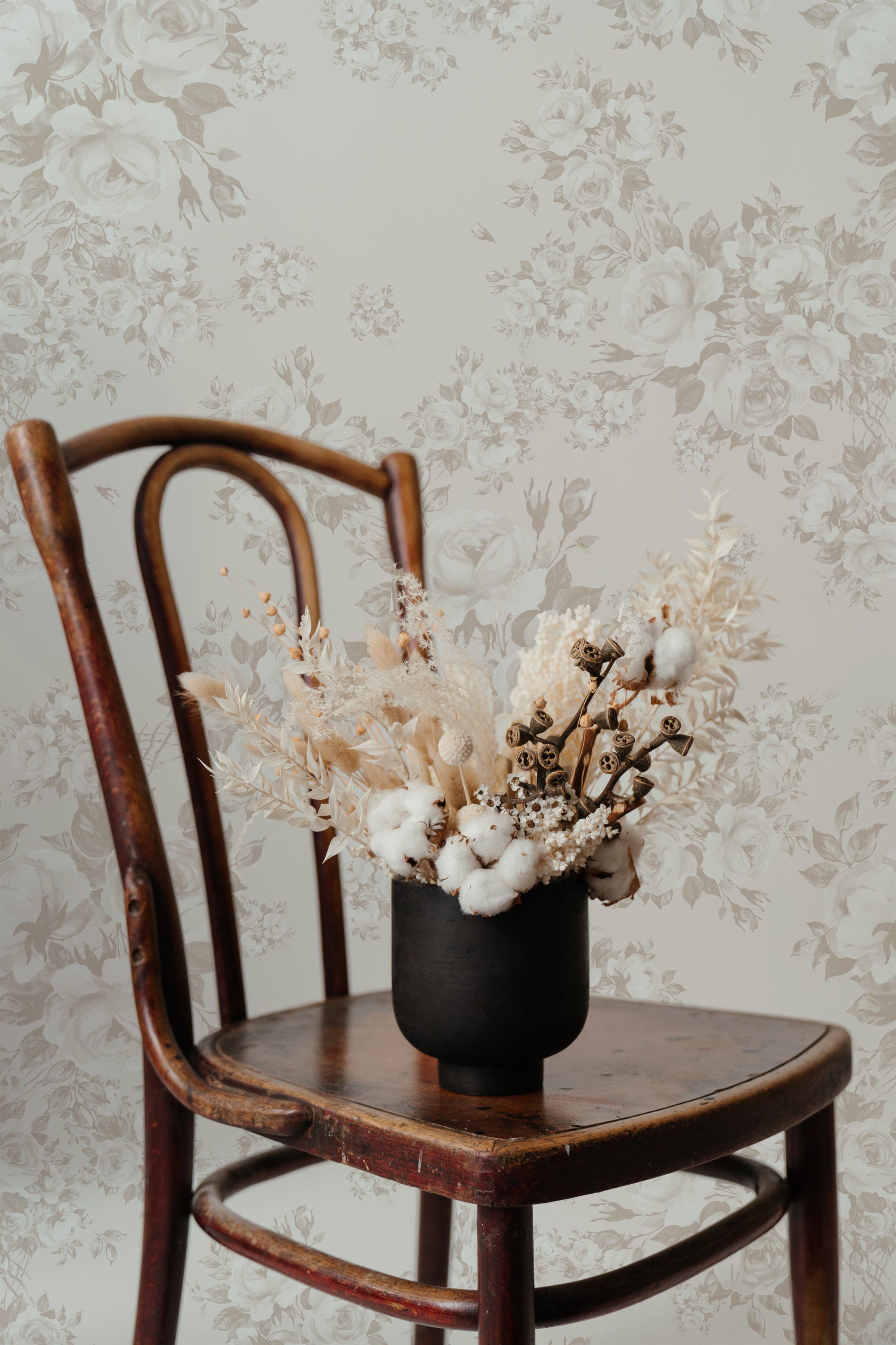 An aged wooden chair set against a wall covered in 'Rose Bouquet Wallpaper,' which showcases a detailed pattern of roses and foliage in muted grey on a light background. Atop the chair is a black vase filled with a mix of dried flowers and cotton branches, complementing the vintage aesthetic of the setting.