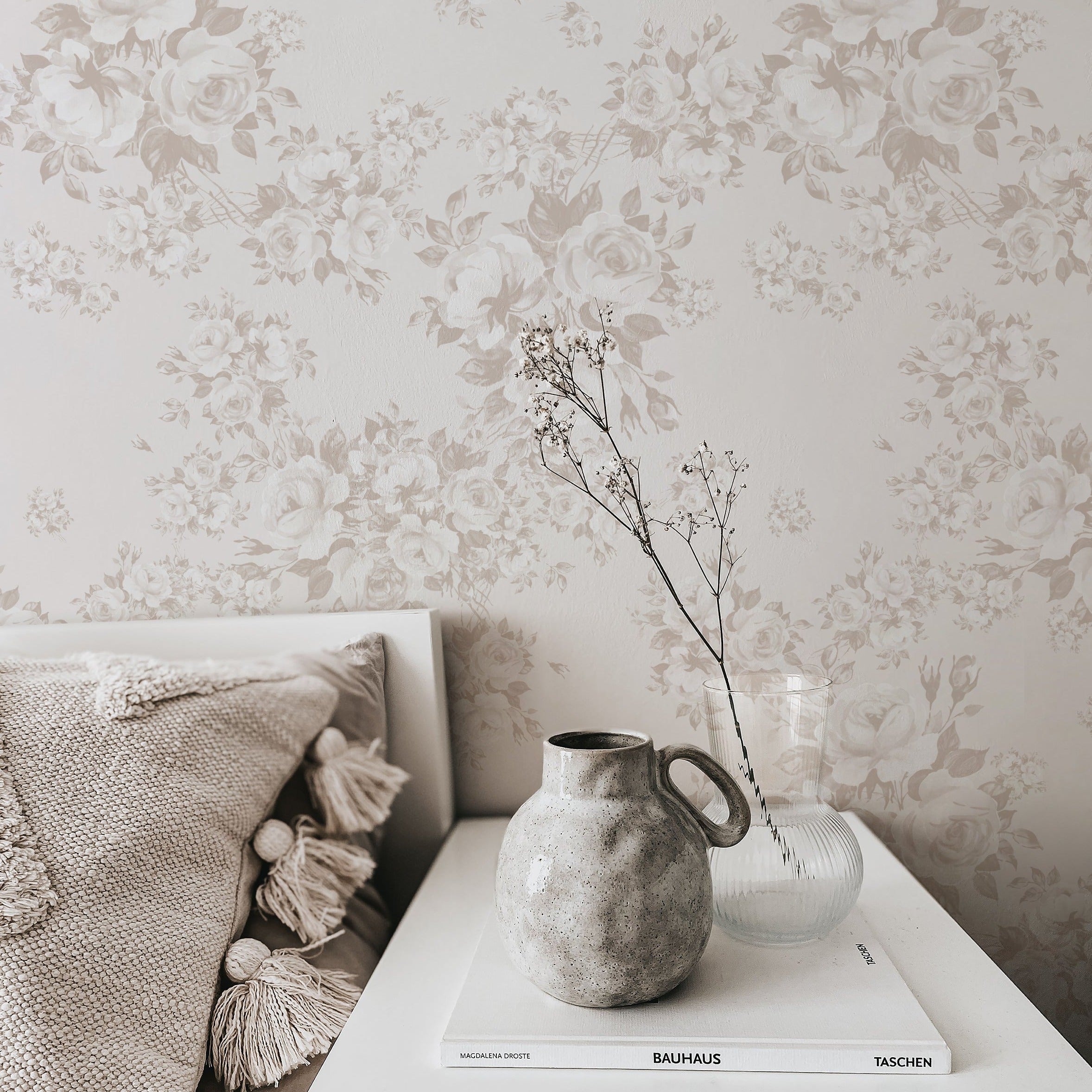 A cozy room corner featuring the 'Rose Bouquet Wallpaper' on the wall, providing a delicate backdrop to a simple home decor scene with a textured cushion, a rustic grey pottery jug, and a clear glass vase with a few sprigs of dried plants, placed on a stack of art books on a white side table.