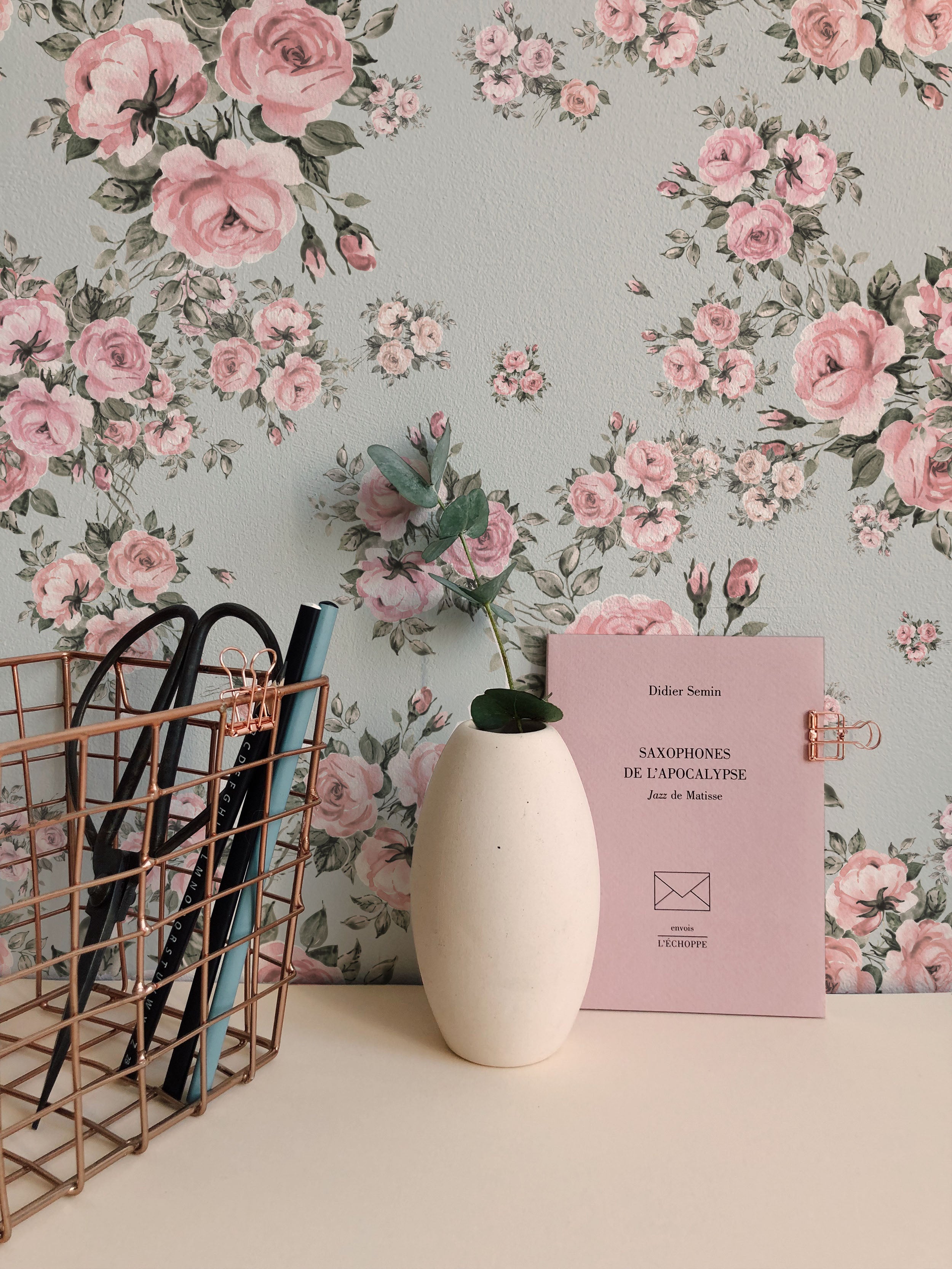 A close-up view of Rose Bouquet Wallpaper II featuring pink roses and green leaves on a light blue background. The setting includes a white vase, books, and a wire basket on a desk.