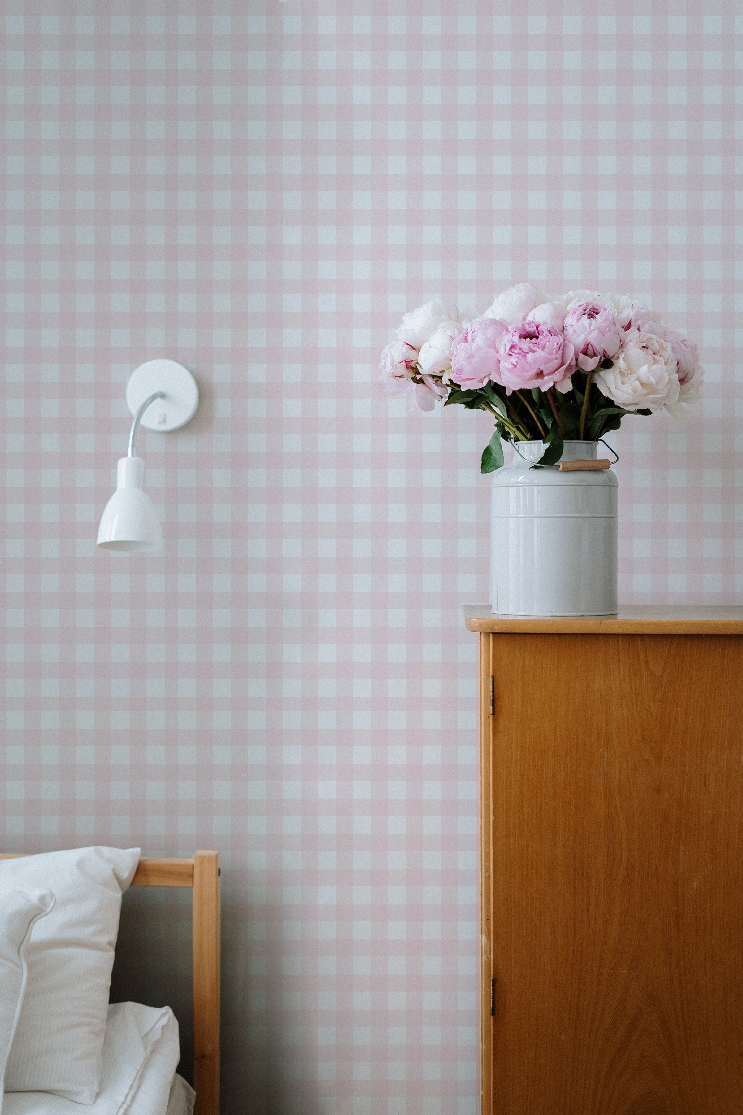 A cozy bedroom corner featuring Gingham Wallpaper in soft pink and white checkered pattern. The wooden bed frame, white pillows, and a wooden dresser with a vase of fresh pink and white flowers complement the wallpaper