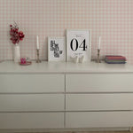 A stylish dresser setup against a wall covered with Gingham Wallpaper in a soft pink and white checkered pattern. Decorative items on the dresser include framed quotes, candles, a stack of books, and a pink flower arrangement