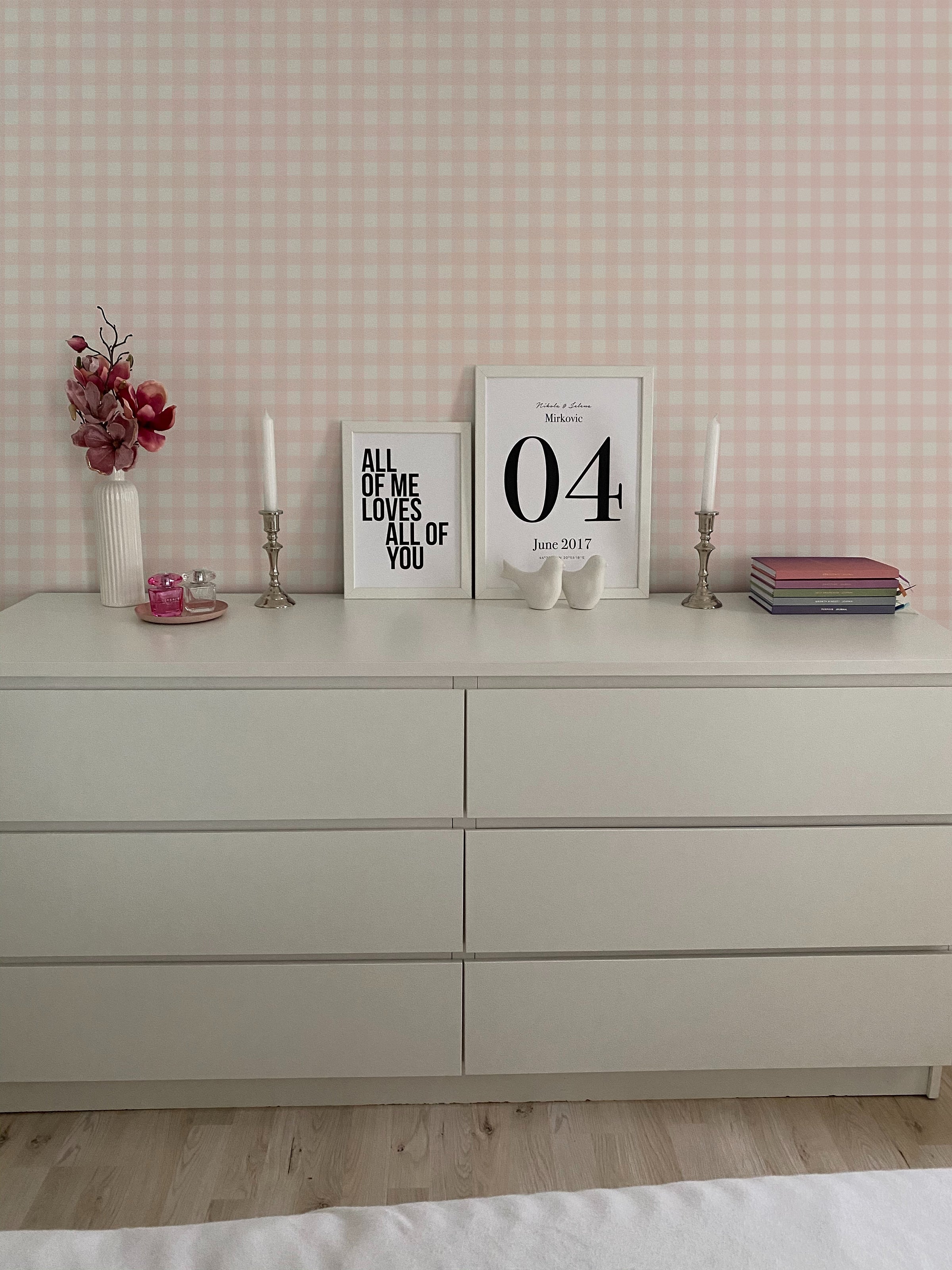 A stylish dresser setup against a wall covered with Gingham Wallpaper in a soft pink and white checkered pattern. Decorative items on the dresser include framed quotes, candles, a stack of books, and a pink flower arrangement