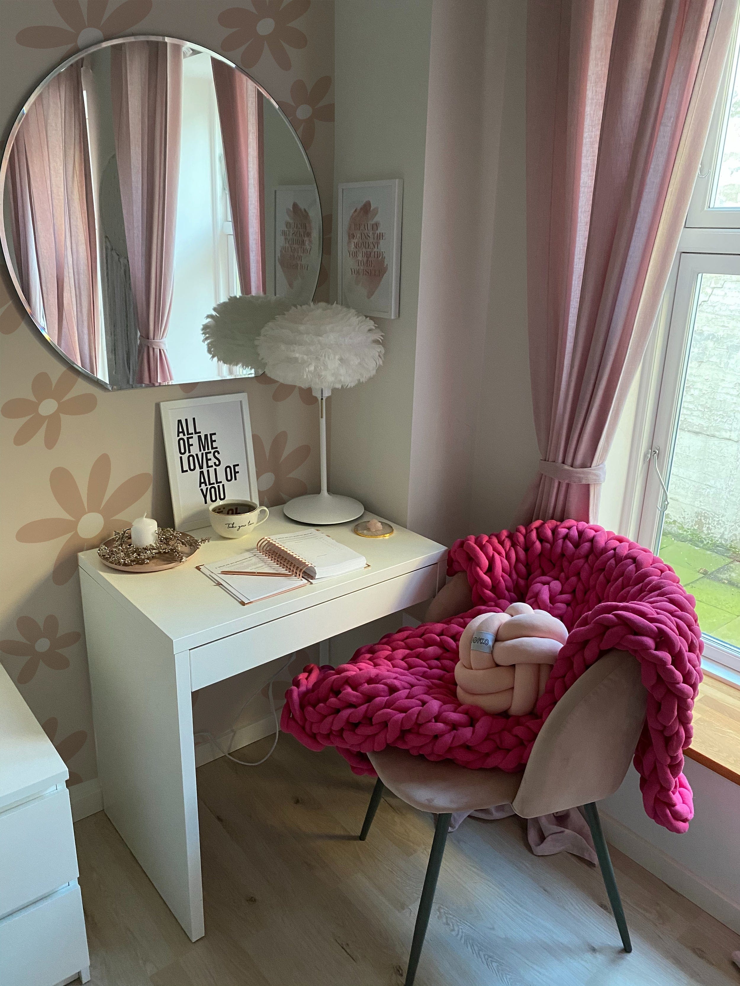 A cozy nook with a white desk and a chair draped with a chunky pink blanket. The wall is adorned with Simple Mauve Floral Wallpaper, complementing the pink curtains and creating a warm, feminine atmosphere. A round mirror and inspirational quotes complete the look.