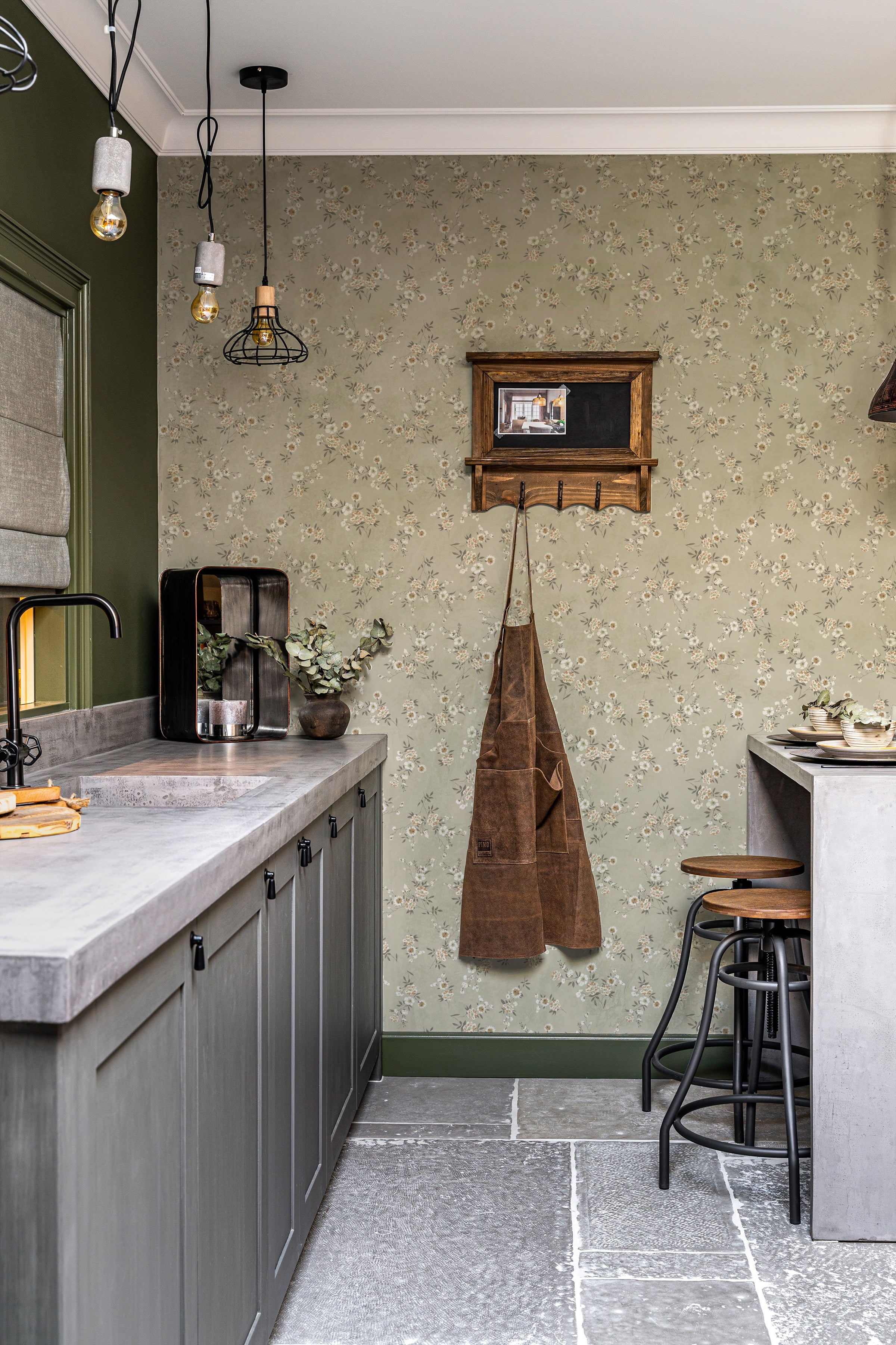 A stylish kitchen featuring the Classic Floral Wallpaper with a muted olive green base and a delicate pattern of white and tan flowers. The wallpaper adds a touch of vintage charm, complemented by the slate gray cabinetry, a concrete countertop, and industrial-style pendant lights. An apron hangs on a wooden hook, adding a rustic touch to the sophisticated decor.