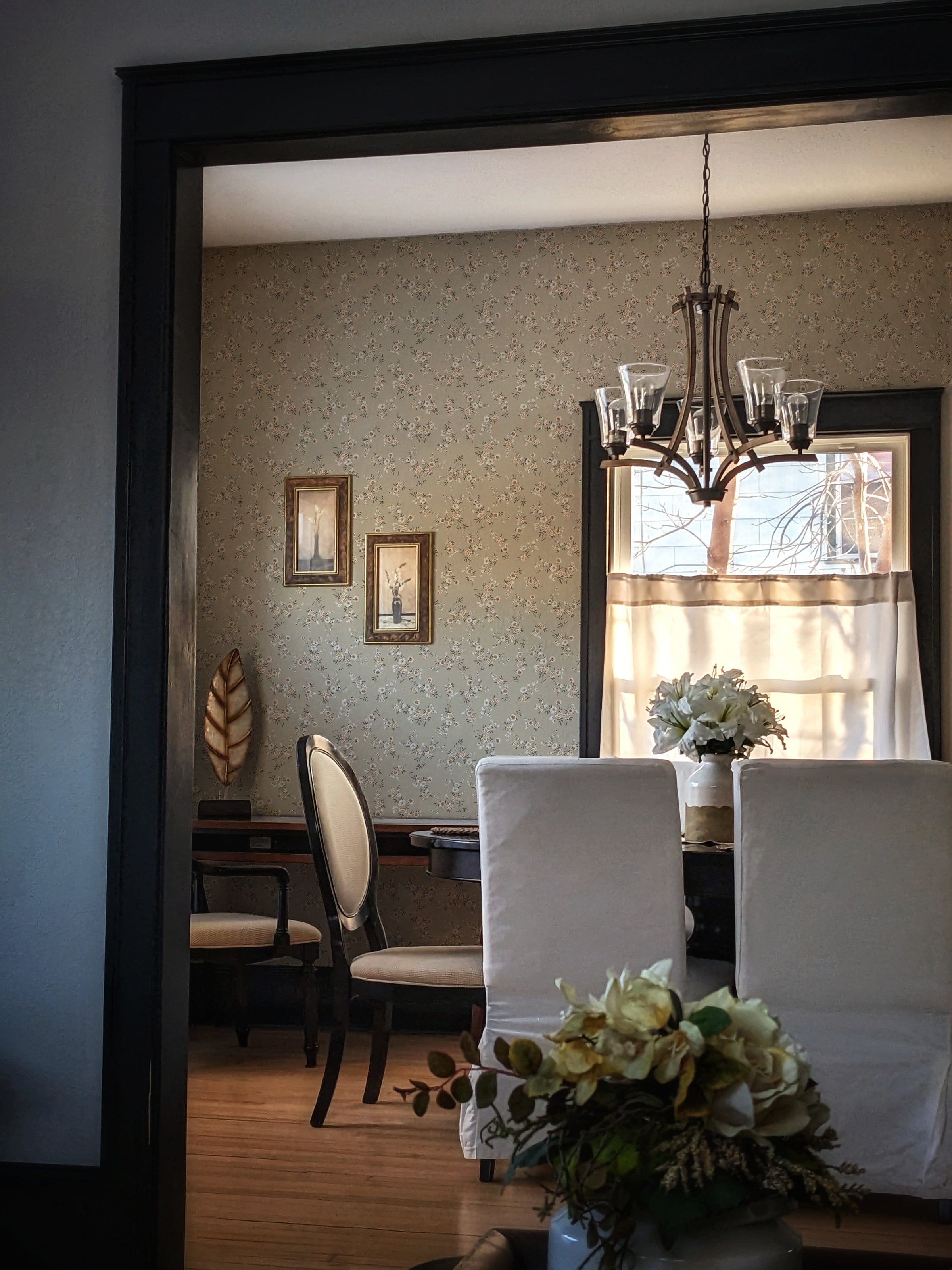 A view into a dining room through a dark frame, showcasing the Classic Floral Wallpaper adorned with an elegant floral design. The room is bathed in warm sunlight, highlighting the antique chandelier and creating a cozy atmosphere. White slipcovered chairs and a table arrangement with flowers complete the inviting scene.
