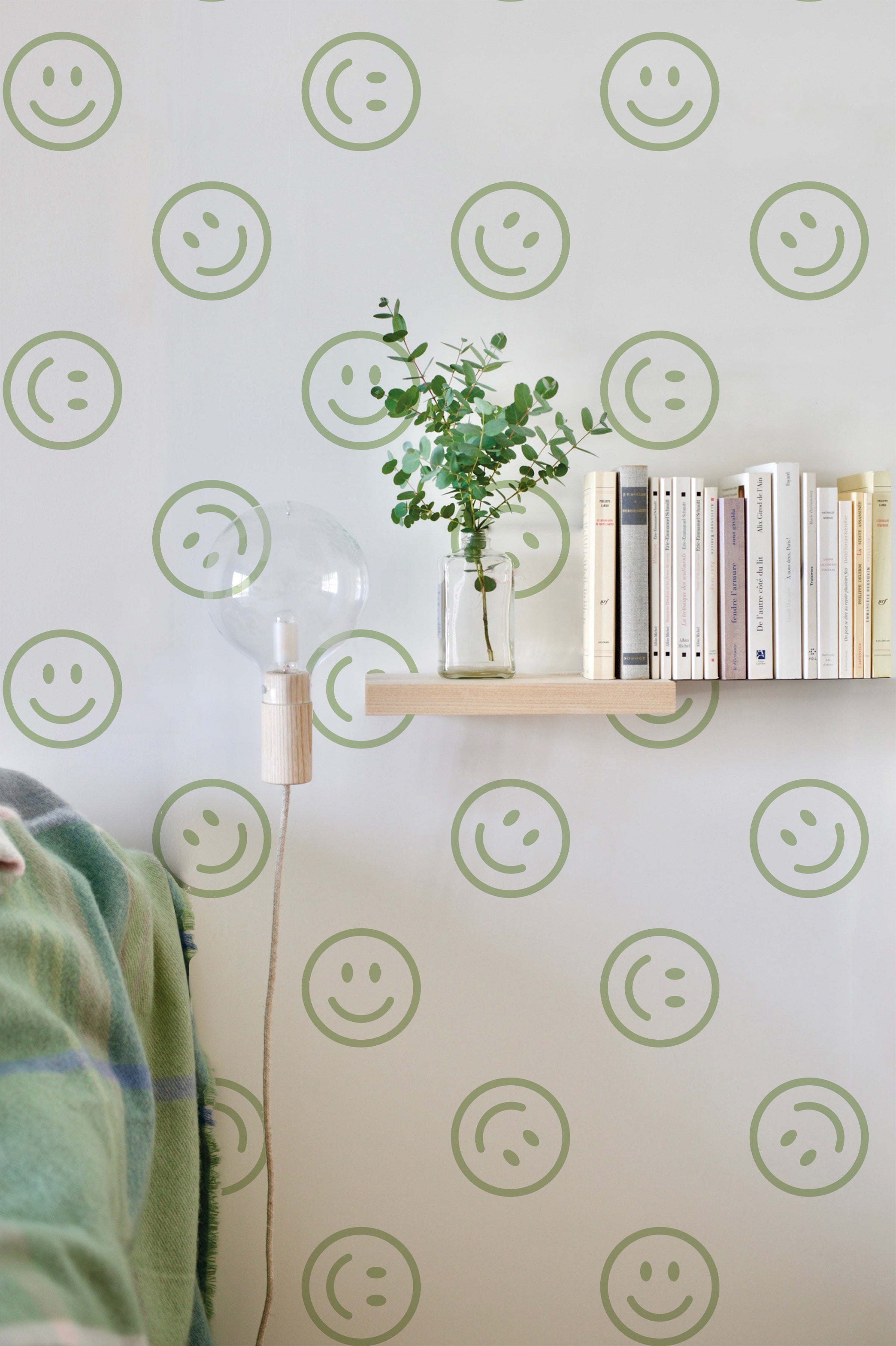 A modern and cheerful room with Smiley Wallpaper featuring green smiley faces on a light background. The decor includes a wooden shelf with books, a vase with greenery, and a unique light fixture.