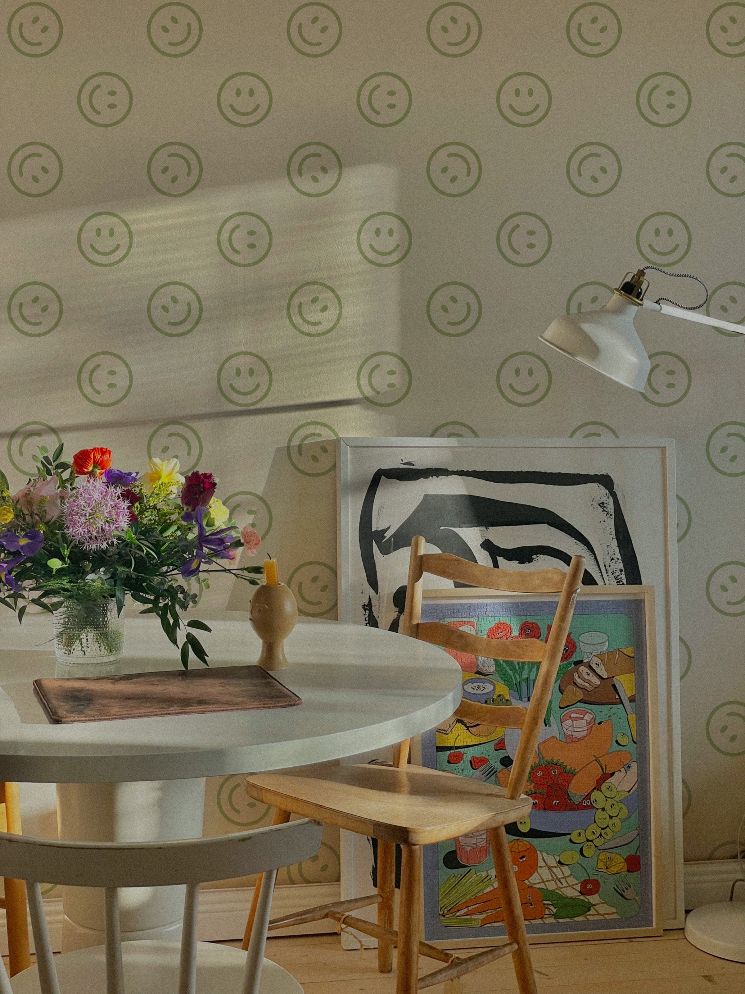 A cozy dining area featuring Smiley Wallpaper with green smiley faces on a light background. The space is decorated with a round white table, wooden chairs, colorful flowers, and framed artwork.