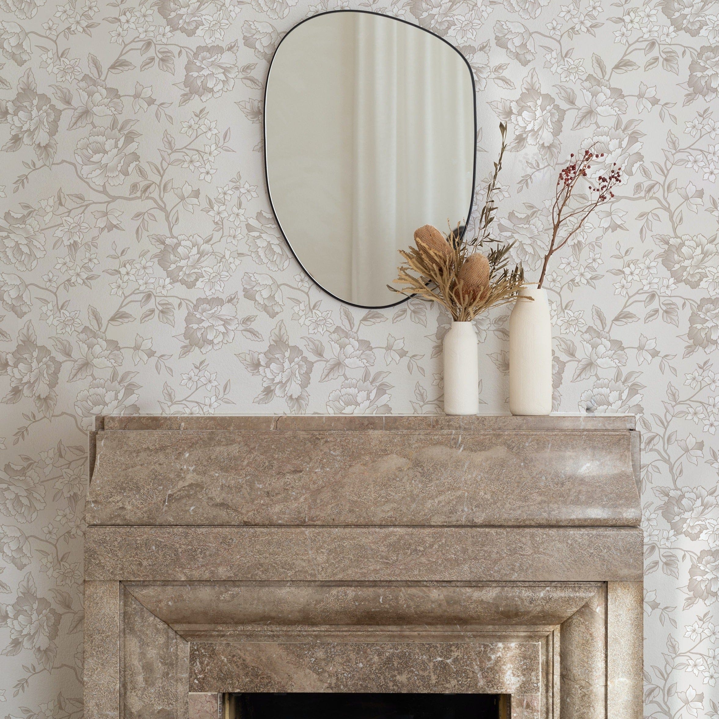 An elegant living room corner with the Ornamental Garden Wallpaper enhancing the wall behind a marble fireplace. The wallpaper's intricate floral pattern adds a luxurious touch, complemented by decorative vases and dried flowers atop the mantelpiece, under a simple round mirror.