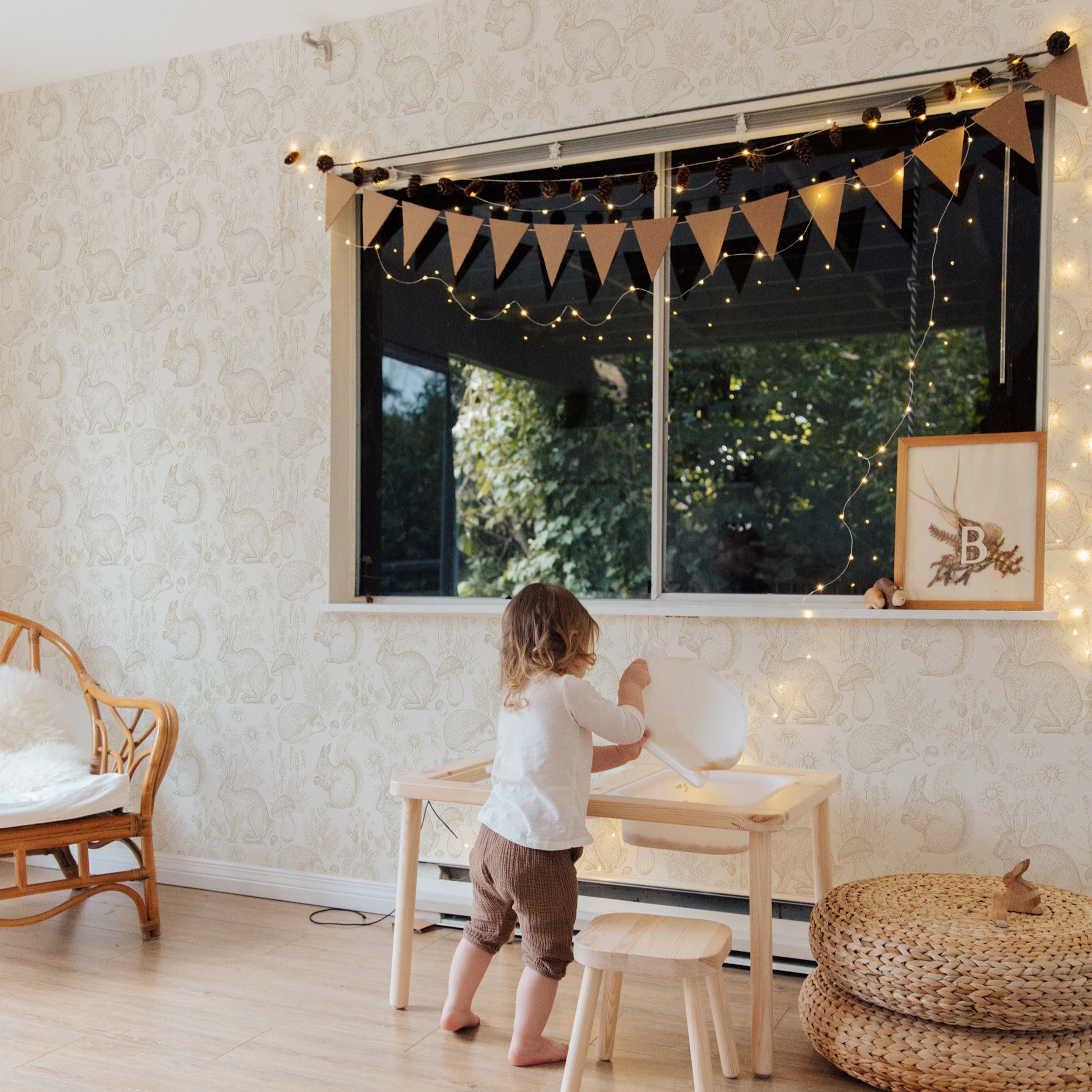 A child's play area against a backdrop of "Woodland Creatures Wallpaper" with playful sketches of rabbits and squirrels. The room features a small wooden table and stools, with a child in a white top and brown pants standing at the window, which is framed with soft drapes and whimsical string lights, creating a magical and kid-friendly environment.