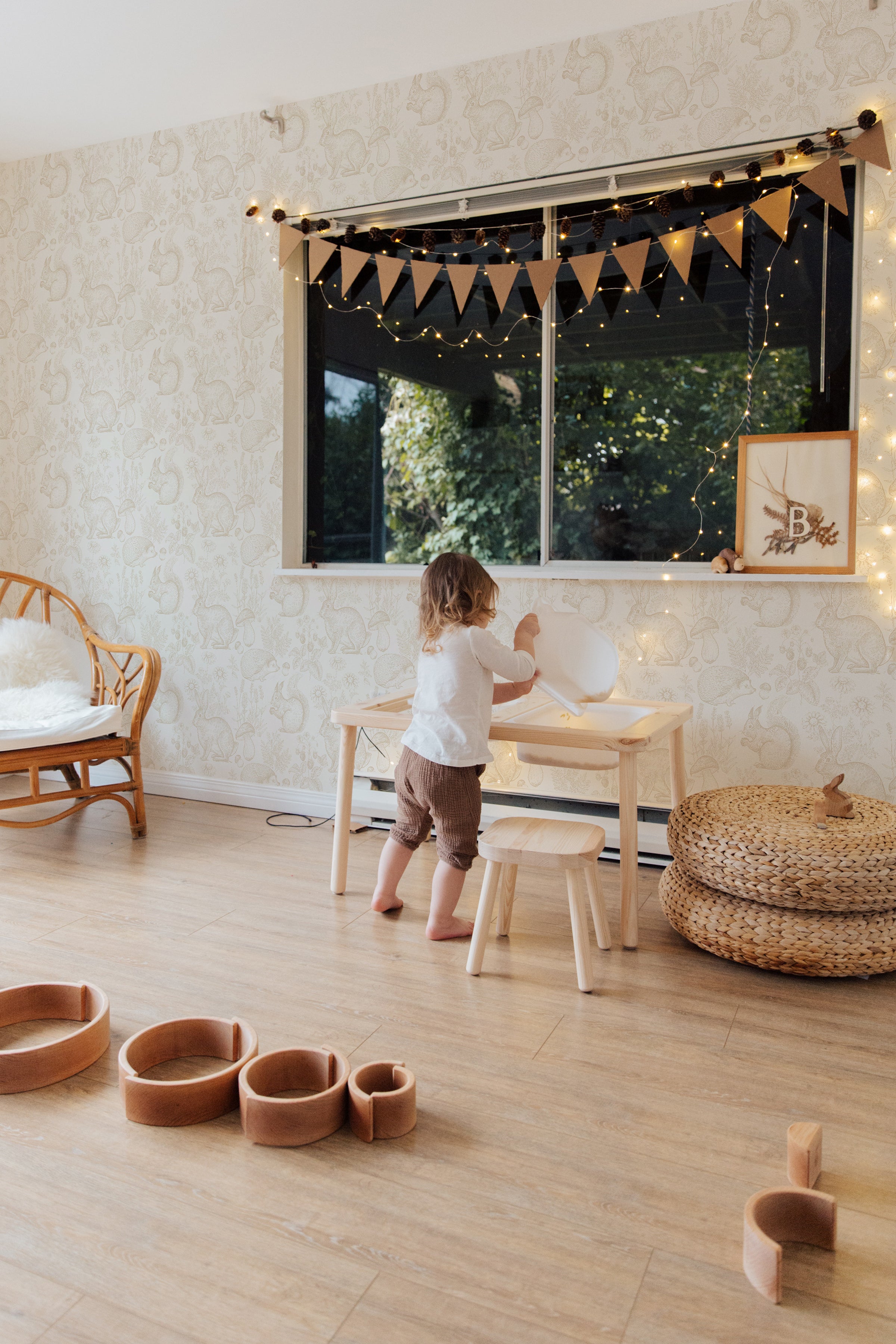 A child's play area against a backdrop of "Woodland Creatures Wallpaper" with playful sketches of rabbits and squirrels. The room features a small wooden table and stools, with a child in a white top and brown pants standing at the window, which is framed with soft drapes and whimsical string lights, creating a magical and kid-friendly environment.