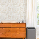 A cozy room corner featuring a mid-century modern wooden dresser against a charming wallpaper adorned with sketched illustrations of woodland creatures like rabbits, squirrels, and hedgehogs amidst flora. A changing pad rests on top of the dresser beside a bottle of lotion and wipes, with a sleek grey diaper pail standing to the side, illustrating a functional and stylish nursery space.