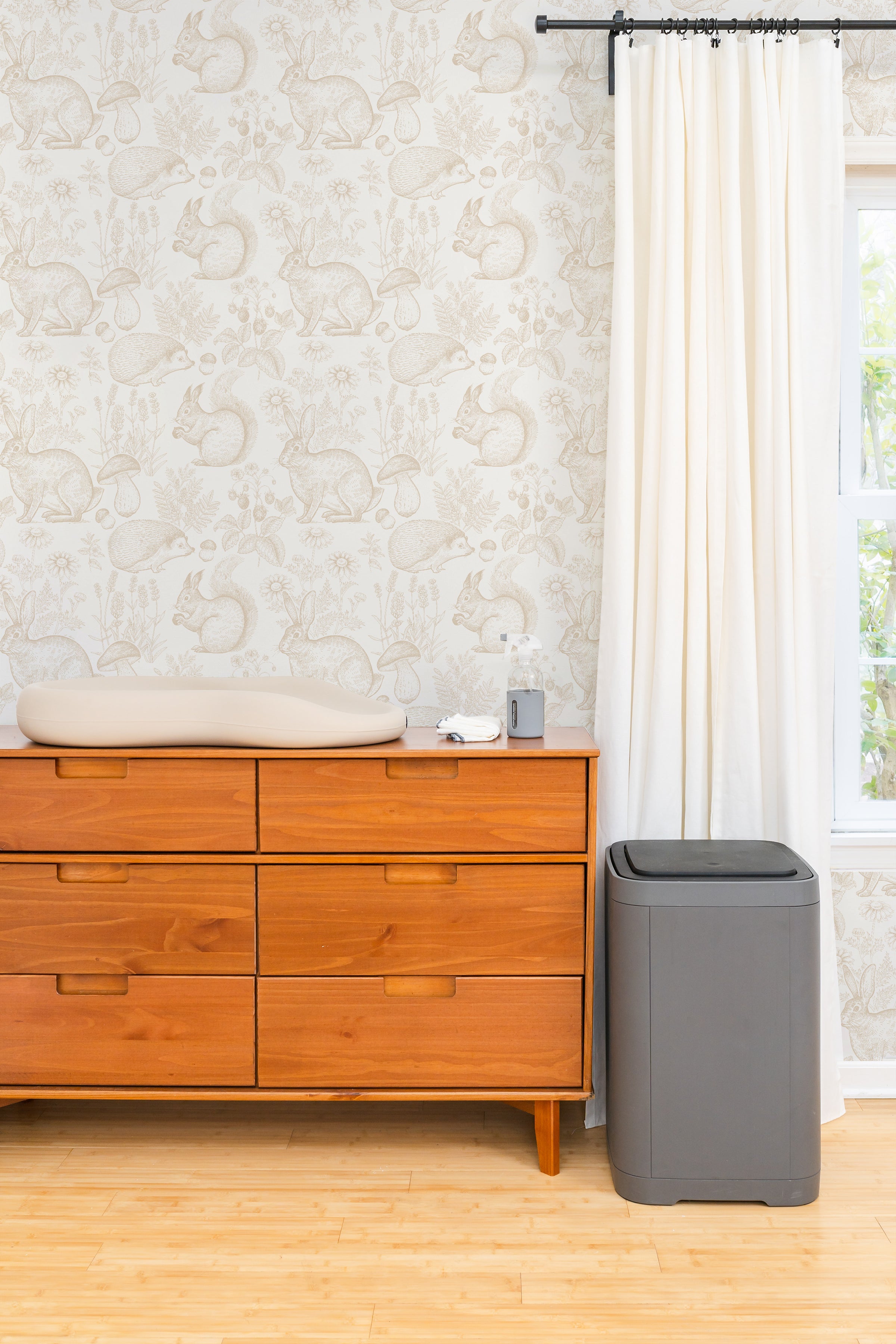 A cozy room corner featuring a mid-century modern wooden dresser against a charming wallpaper adorned with sketched illustrations of woodland creatures like rabbits, squirrels, and hedgehogs amidst flora. A changing pad rests on top of the dresser beside a bottle of lotion and wipes, with a sleek grey diaper pail standing to the side, illustrating a functional and stylish nursery space.