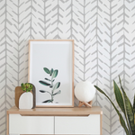 A styled room with the Hand Painted Chevron Arrow Wallpaper adorning the wall, complemented by a modern dresser, a framed botanical print, a wooden heart sculpture, and a spherical textured lamp, creating a harmonious blend of contemporary and handmade aesthetics