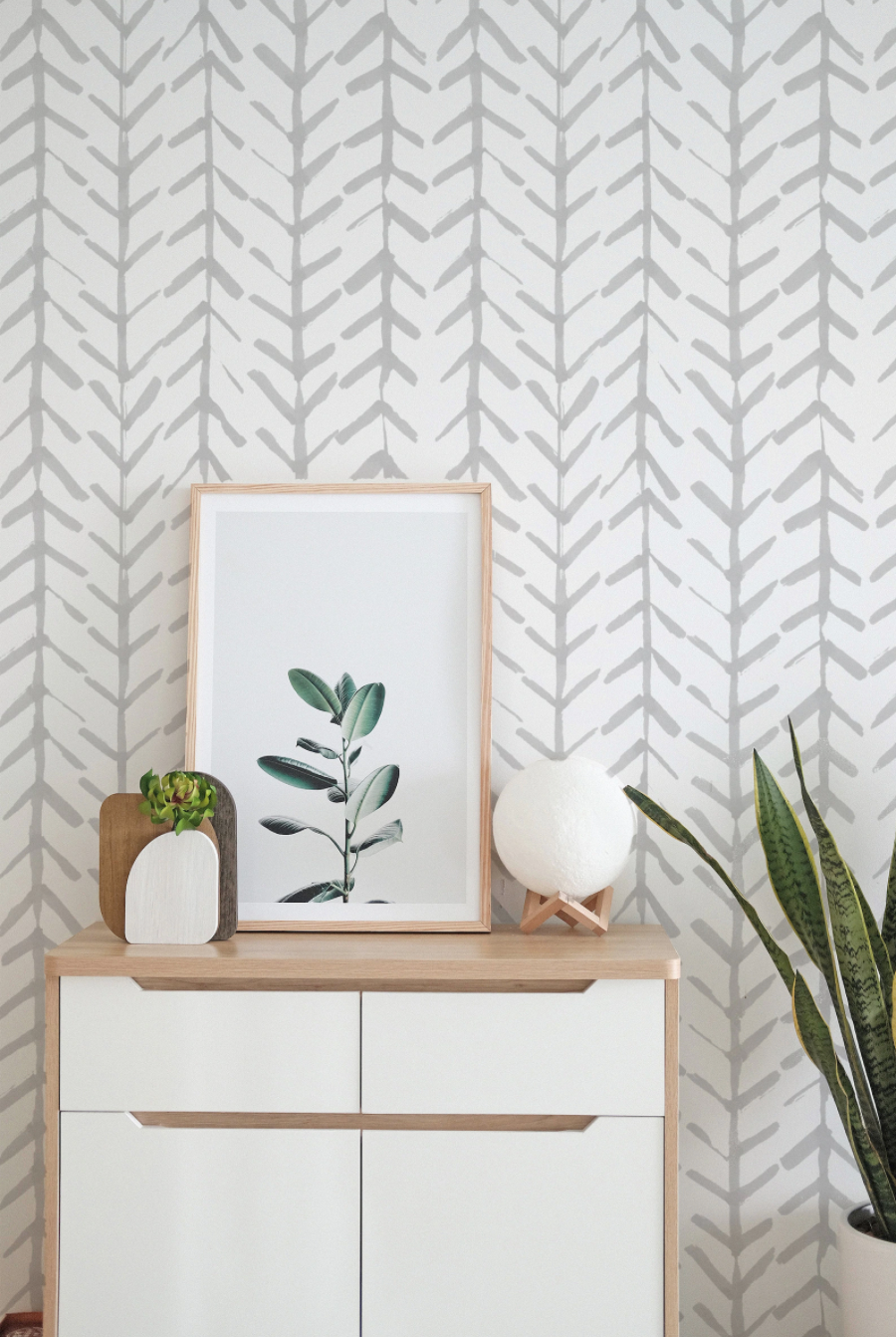 A styled room with the Hand Painted Chevron Arrow Wallpaper adorning the wall, complemented by a modern dresser, a framed botanical print, a wooden heart sculpture, and a spherical textured lamp, creating a harmonious blend of contemporary and handmade aesthetics