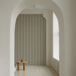 A view through an arched doorway showing a wall adorned with 'Burlap Striped Wallpaper', creating a sense of depth and texture with its muted stripes, paired with a minimalist wooden stool