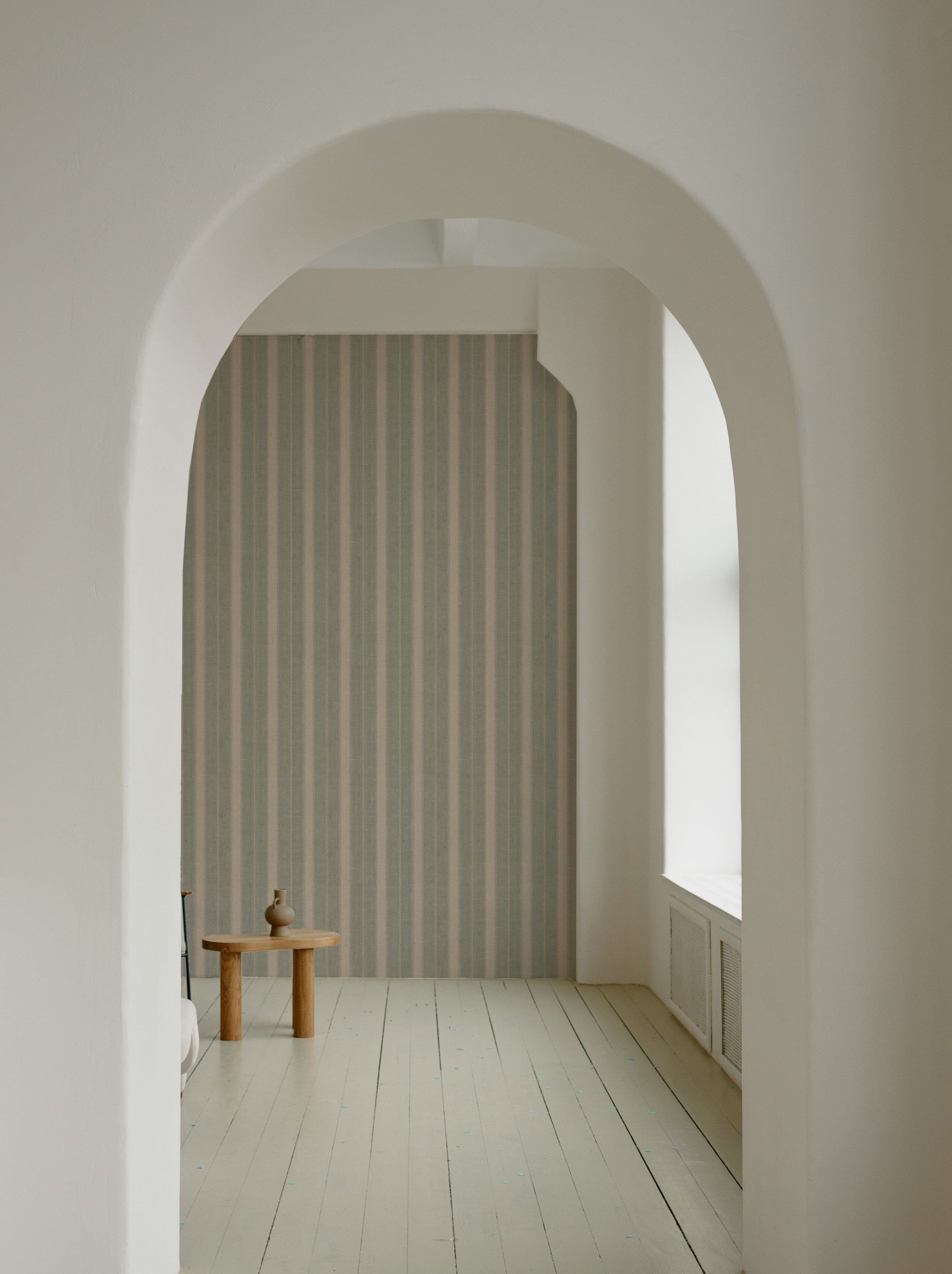 A view through an arched doorway showing a wall adorned with 'Burlap Striped Wallpaper', creating a sense of depth and texture with its muted stripes, paired with a minimalist wooden stool