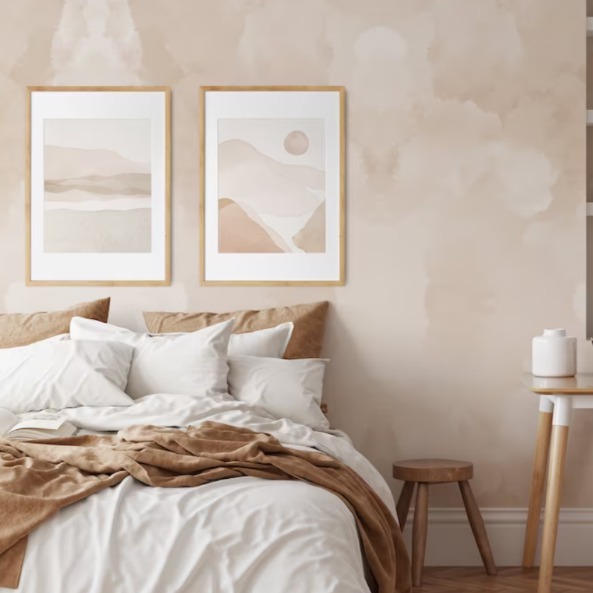 A cozy bedroom featuring two large framed abstract prints above the bed. The prints are in soft earth tones, with one depicting a minimalist desert landscape and the other an abstract rendition of sun and hills. The wall behind them has a watercolor mural in a matching beige hue.