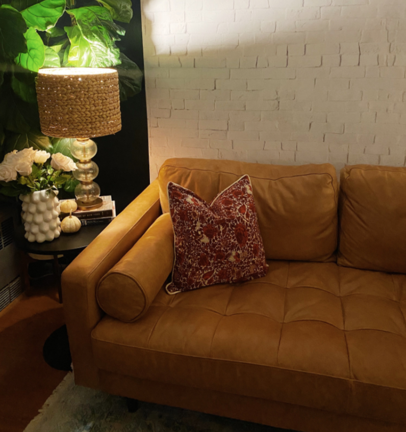 The corner of a stylish living room, where the Realistic White Brick Wallpaper adds an element of loft-like sophistication. The wallpaper is paired with a tan leather couch and exotic patterned cushions, while a decorative lamp and plant arrangement complete the look, creating a harmonious blend of urban texture and home comfort.