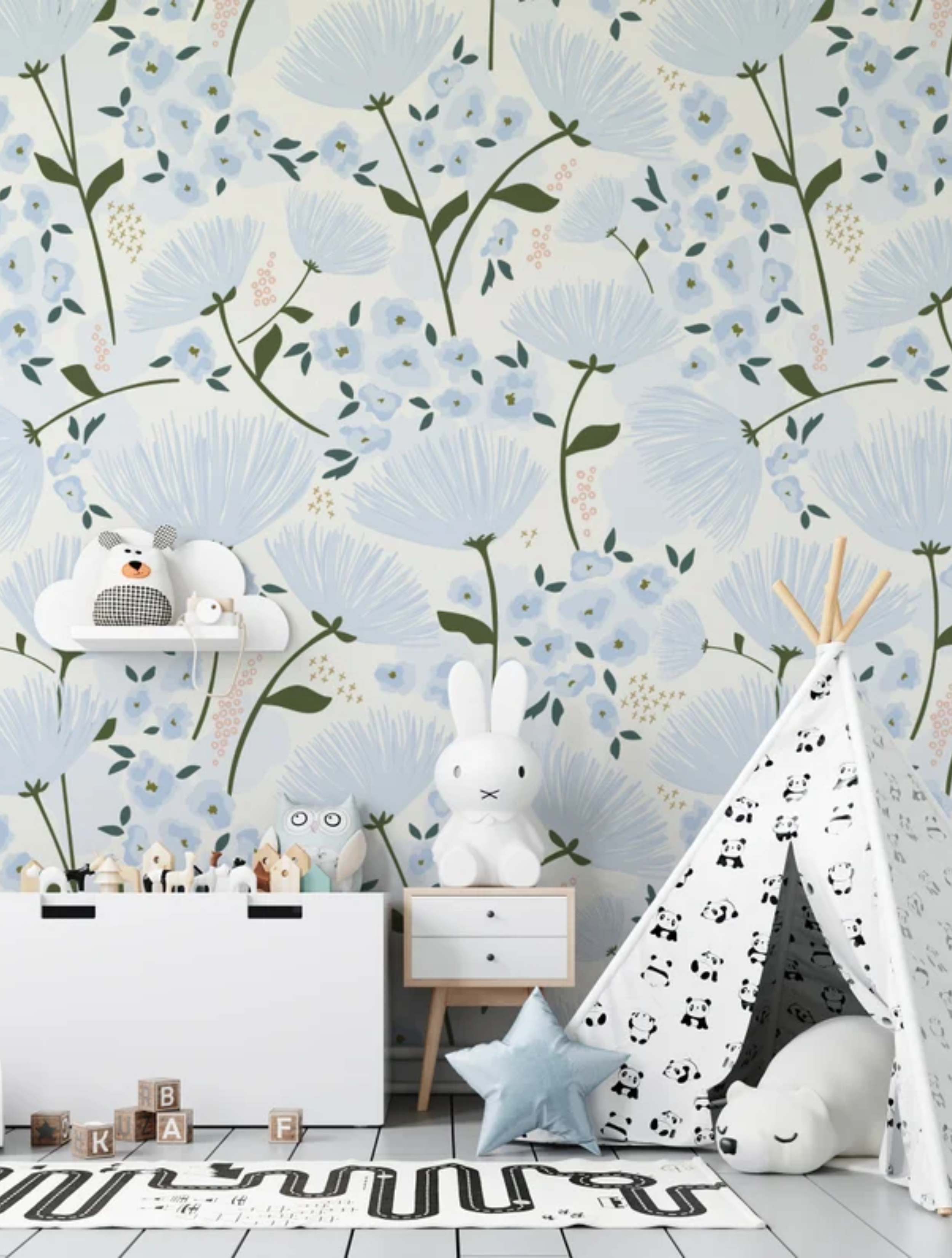 A charming children's play area is brightened by the "Floral Love Wallpaper," which is adorned with delicate blue flowers and soft green foliage on a light background. The space is playfully styled with a child's teepee, a toy chest, and whimsical stuffed animals, enhancing the gentle and joyful atmosphere created by the wallpaper.