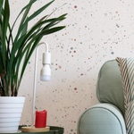 A cozy corner of a room adorned with Ink Spatter Wallpaper, enhancing the area with its joyful splatter pattern. Nearby, a modern white lamp and a green upholstered chair beside a potted plant complement the wallpaper's colorful accents, creating a cheerful space.