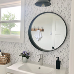 Modern bathroom adorned with Dainty Black Floral Wallpaper, showcasing a circular black-framed mirror above a white sink with a chrome faucet.
