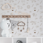 A cozy nursery corner showcasing Safari Jungle Wallpaper with a cute array of hand-drawn-style safari animals on a soft beige background. The scene includes a white storage unit with wicker baskets, a playful bear lamp, children’s accessories, and a wooden coat rack adorned with a charming cloud-shaped clock and baby essentials.