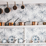 A kitchen scene featuring Line Minimal Floral Leaf wallpaper, with a sketched design of thin-lined floral leaves in blue on a white background. Above a white shelf holding decorative plates and mugs, copper pots and pans hang, adding a rustic charm against the contemporary floral backdrop.