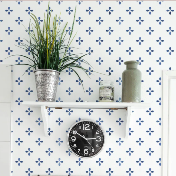 A wall adorned with the Azulejo - True Blue Wallpaper, complemented by a simple white shelf displaying a potted plant, a ceramic vase, and a vintage-style black wall clock. The blue patterned wallpaper adds a touch of Mediterranean charm to the otherwise minimalist decor.
