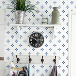 A wall adorned with the Azulejo - True Blue Wallpaper, complemented by a simple white shelf displaying a potted plant, a ceramic vase, and a vintage-style black wall clock. The blue patterned wallpaper adds a touch of Mediterranean charm to the otherwise minimalist decor.